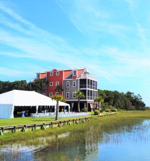 A colorful house by a calm waterway, with a large white tent set up on the lawn for an event. Clear blue sky above.