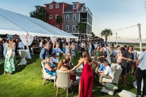 A lively outdoor event with guests mingling under a tent, surrounded by greenery and colorful buildings in the background.