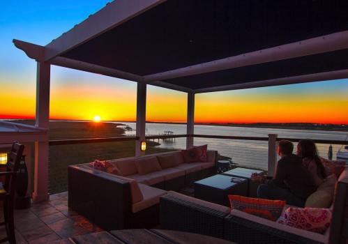 A couple sits on a patio, watching a vibrant sunset over a calm waterway. Cozy seating and a serene atmosphere.