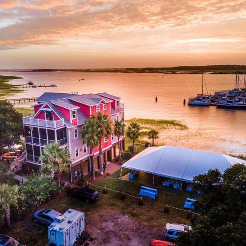 Aerial view of a colorful house by the water at sunset, with boats and a large tent in the foreground.