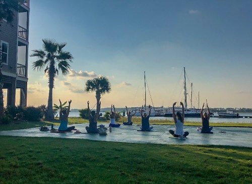 A group of people practicing yoga outdoors by the water, with palm trees and boats in the background.