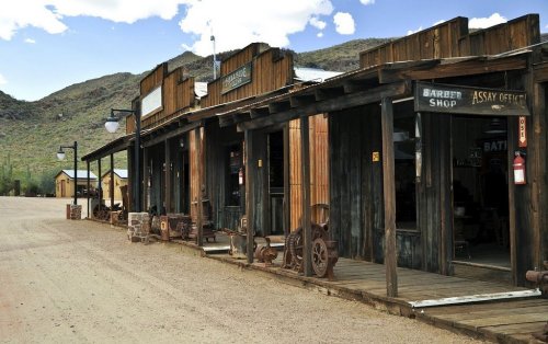 A rustic old western town with wooden storefronts and a dirt road, set against a mountainous backdrop.