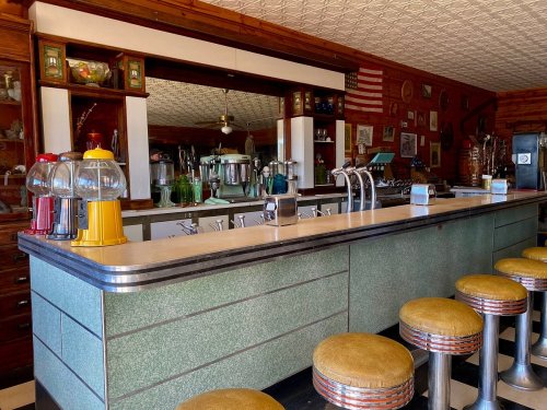 A vintage diner counter with green and chrome details, yellow and red soda dispensers, and round stools.