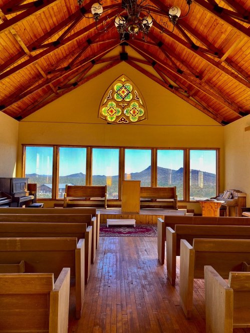 Interior of a chapel with wooden pews, a podium, and a stained glass window, overlooking mountains through large windows.