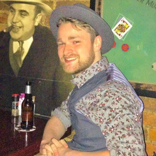 A young man in a hat and patterned shirt smiles at the camera, sitting at a bar with a vintage backdrop.
