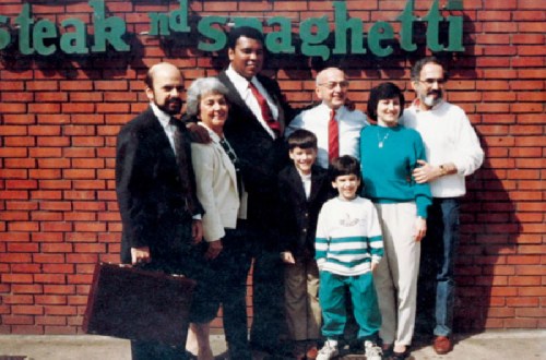 A group of seven people, including children and adults, pose together in front of a restaurant sign that reads "Steak and Spaghetti."