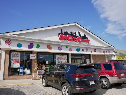 Exterior of All City Candy store with colorful decorations and a clear blue sky. Cars parked in front.