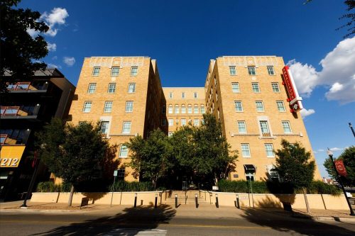 A large, tan brick building with multiple floors, flanked by trees and a clear blue sky.