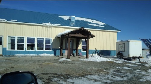 A building with a blue roof and snow on the ground, featuring a covered entrance and a trailer nearby.