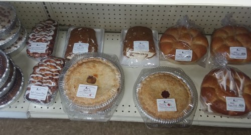 A display of various baked goods, including pies, cakes, and rolls, all packaged with labels on a shelf.