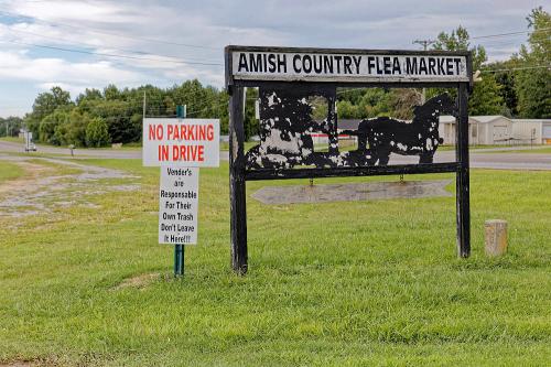 Sign for Amish Country Flea Market with a no parking notice and guidelines for vendors on trash disposal. Green grass in foreground.