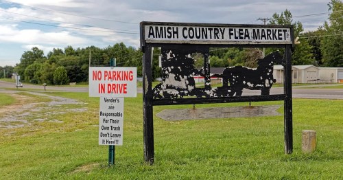 Sign for "Amish Country Flea Market" with a warning about parking and vendor responsibilities for trash. Green grass and trees in background.