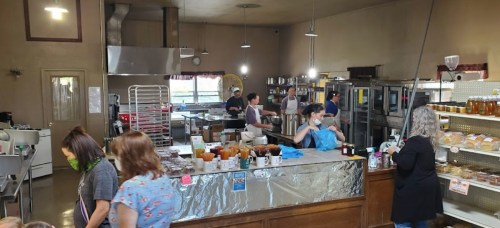 A busy kitchen with staff preparing food, shelves of jars, and equipment in a warm, inviting atmosphere.