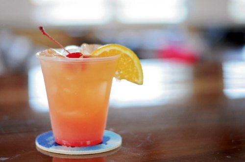 A colorful cocktail in a clear cup, garnished with a cherry and an orange slice, on a wooden table.