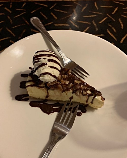 A slice of cheesecake drizzled with chocolate sauce, topped with whipped cream, served on a white plate with two forks.