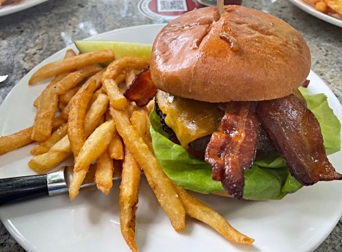 A juicy burger with bacon and cheese on a bun, served with crispy fries and a pickle on a white plate.