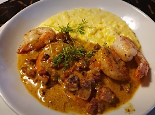 A plate of shrimp in a creamy sauce served with a side of yellow grits, garnished with fresh herbs.