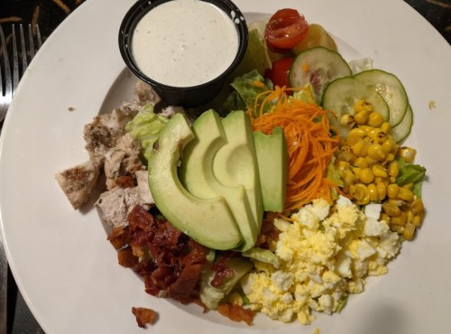 A colorful salad plate with chicken, avocado, corn, cucumbers, tomatoes, carrots, and a side of dressing.