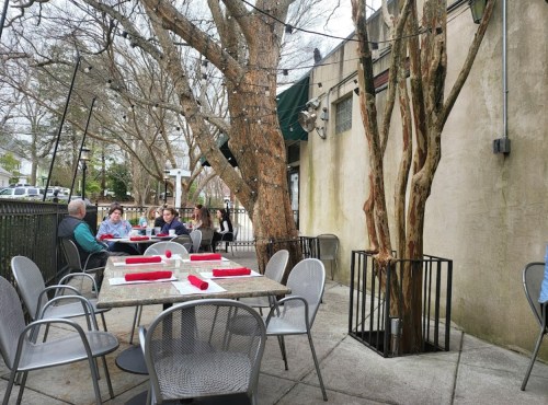 Outdoor dining area with tables, chairs, and trees, featuring diners enjoying a meal under string lights.