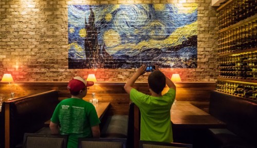 Two children take a photo of a large painting resembling a starry night scene in a cozy restaurant.
