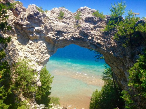 A natural stone arch frames a view of clear blue water and a rocky shoreline, surrounded by lush greenery.