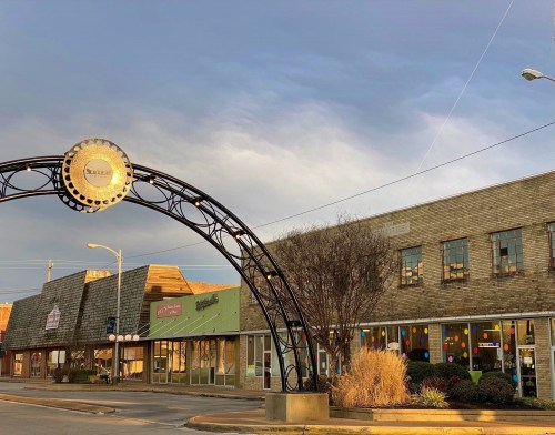 A street view featuring a decorative archway and storefronts with colorful windows under a cloudy sky.