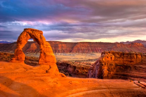 A stunning view of Delicate Arch at sunset, with vibrant orange rock formations and dramatic clouds in the sky.