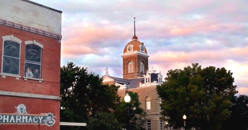 Historic building with a clock tower, surrounded by trees and a pharmacy sign, under a colorful sky at dusk.