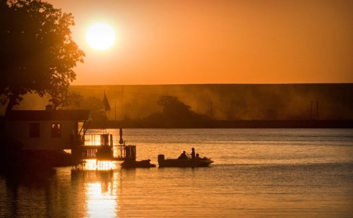 Silhouetted figures on a boat at sunset, with a calm lake and a house by the water reflecting golden hues.