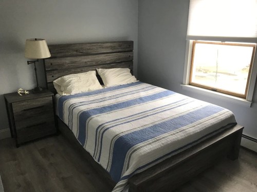A cozy bedroom featuring a wooden bed frame, blue and white striped blanket, nightstand, and a lamp by the window.