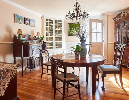 A cozy dining room with a wooden table, chairs, chandelier, and decorative plants, featuring warm wooden floors.