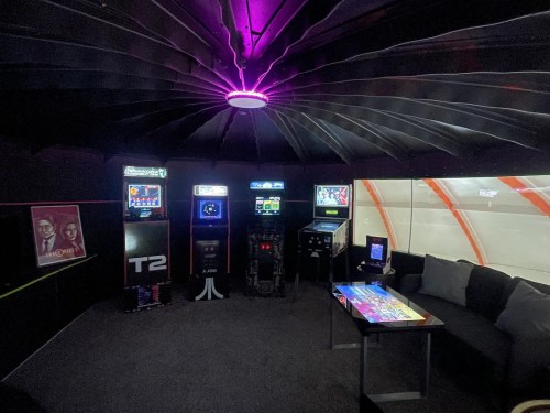 A cozy gaming room featuring retro arcade machines, a modern table, and vibrant purple lighting on the ceiling.