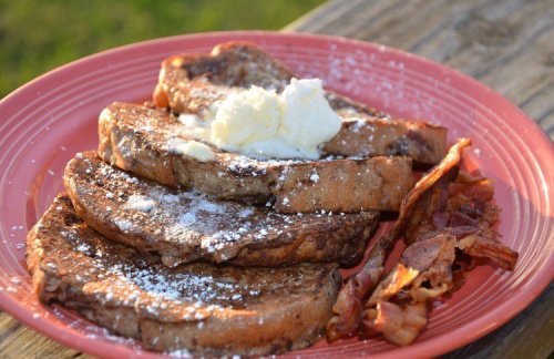A plate of French toast topped with butter and powdered sugar, served with crispy bacon on the side.