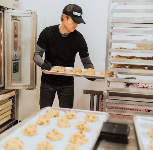 A person in a black shirt and cap places cookie dough on a baking tray in a kitchen.