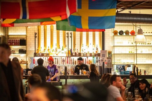 A lively bar scene with flags hanging above, featuring bartenders serving drinks and patrons enjoying their time.