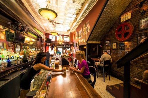 A lively bar scene with patrons, a bartender serving drinks, and vintage decor on the walls.