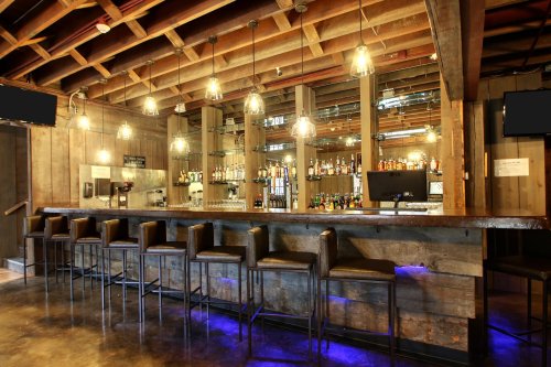 A rustic bar with wooden beams, a long counter, and shelves stocked with various bottles under warm lighting.