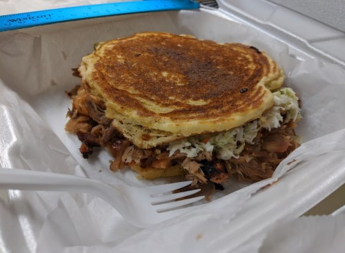 A takeout container with a stacked sandwich made of pulled pork and coleslaw between two golden pancakes.