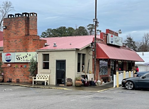 A rustic restaurant with a red roof and brick chimney, featuring outdoor seating and a sign that reads "Myral's Pit Bar."