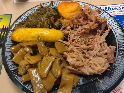 A black plate with pulled pork, cornbread, collard greens, pickles, and green beans.