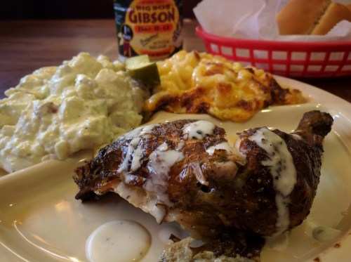 A plate featuring grilled chicken with sauce, creamy macaroni and cheese, potato salad, and a drink in the background.