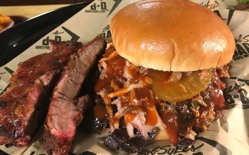 A close-up of a BBQ sandwich with pulled pork, pickles, and sauce, alongside two slices of smoked beef brisket.