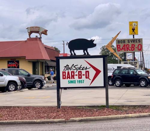 Sign for Bob Sykes Bar-B-Q, featuring a pig silhouette and a vintage-style sign, with a restaurant in the background.