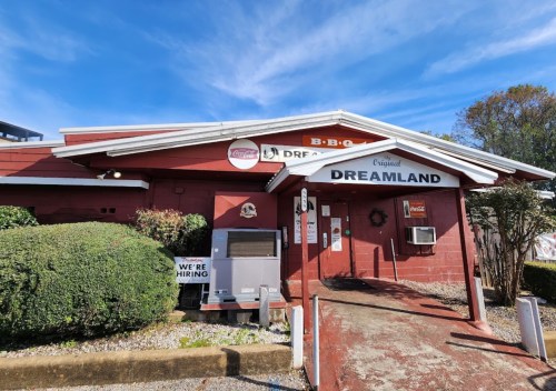 Exterior of Dreamland BBQ restaurant with a red facade, sign, and hiring notice, surrounded by greenery and blue sky.