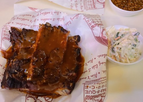 A plate of barbecue ribs covered in sauce, served with coleslaw and baked beans on the side, wrapped in paper.