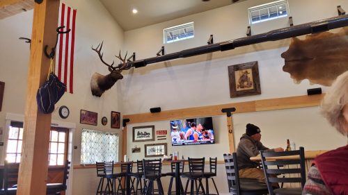 Interior of a rustic restaurant featuring wooden beams, a mounted deer head, and a TV showing a sports event.