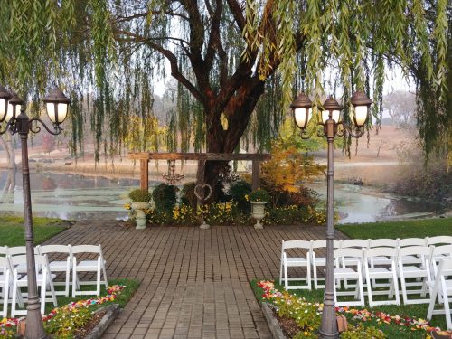 A serene outdoor wedding setup by a pond, featuring white chairs, a wooden arch, and lush greenery.