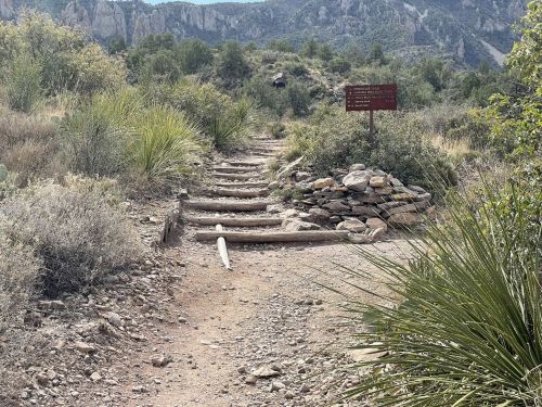 A dirt trail leads through a rocky landscape, with a sign and stone pile visible among shrubs and mountains in the background.
