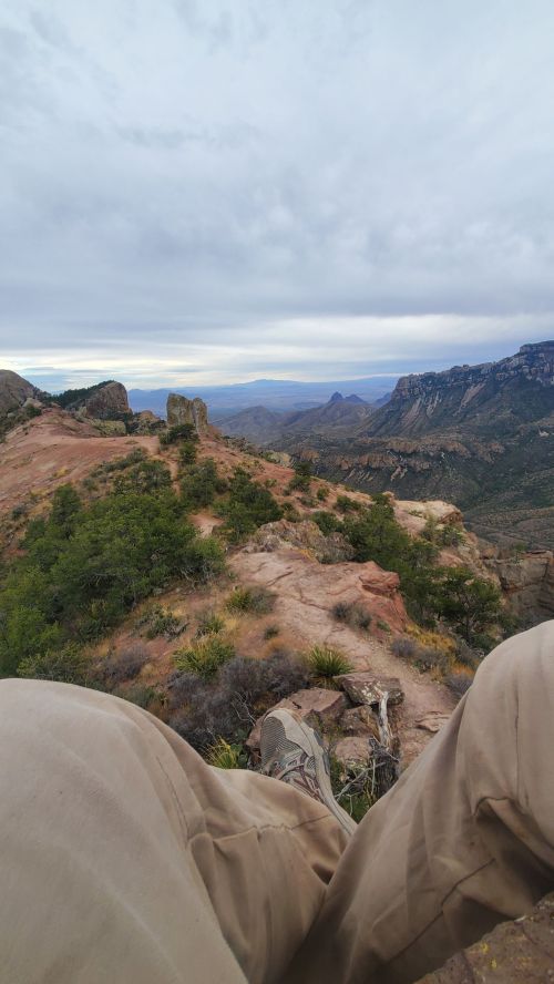 A scenic view from a rocky ledge, overlooking a vast landscape of mountains and valleys under a cloudy sky.