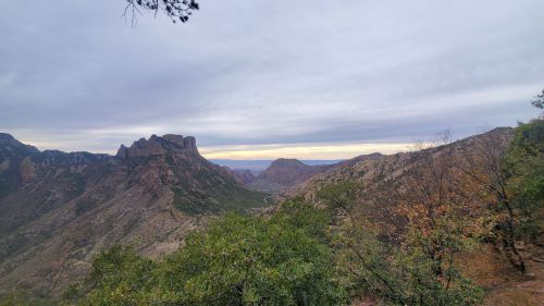 A panoramic view of rugged mountains and valleys under a cloudy sky, with trees in the foreground.